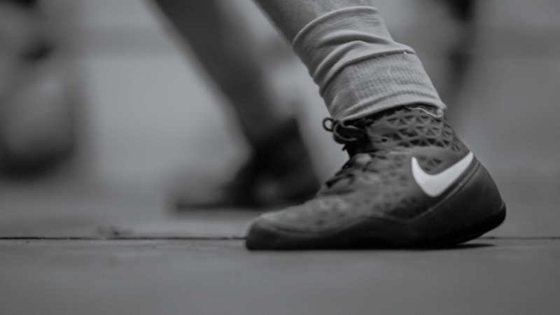 Black Lab Films | Close-up of a person's foot wearing a Nike wrestling shoe with a white sock on a floor surface.