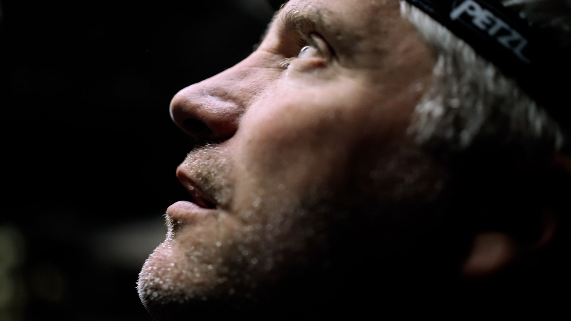 Black Lab Films | A side profile of a person looking upward, illuminated by a light source, wearing a Petzl headlamp in a dark setting.