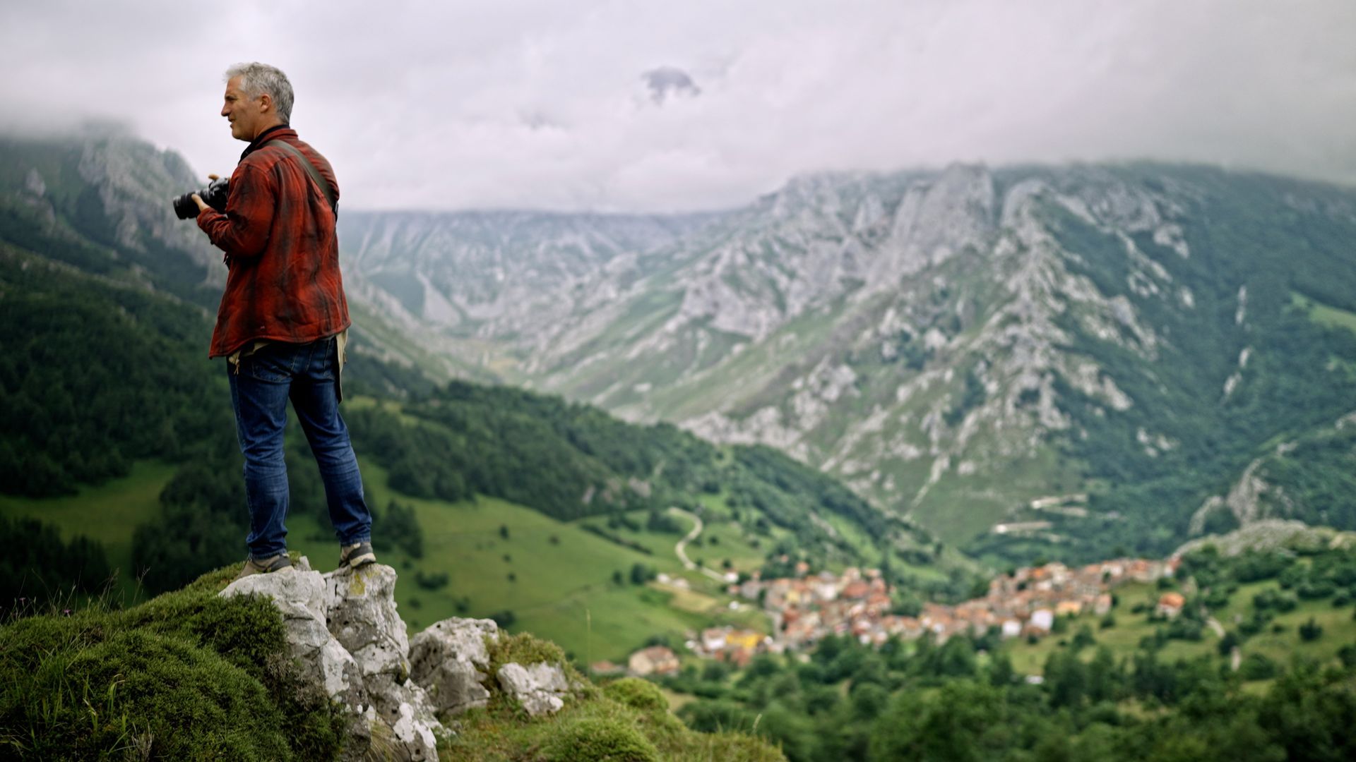 Black Lab Films | A person stands on a rocky outcrop holding a camera, overlooking a lush valley with a village nestled in the mountains.