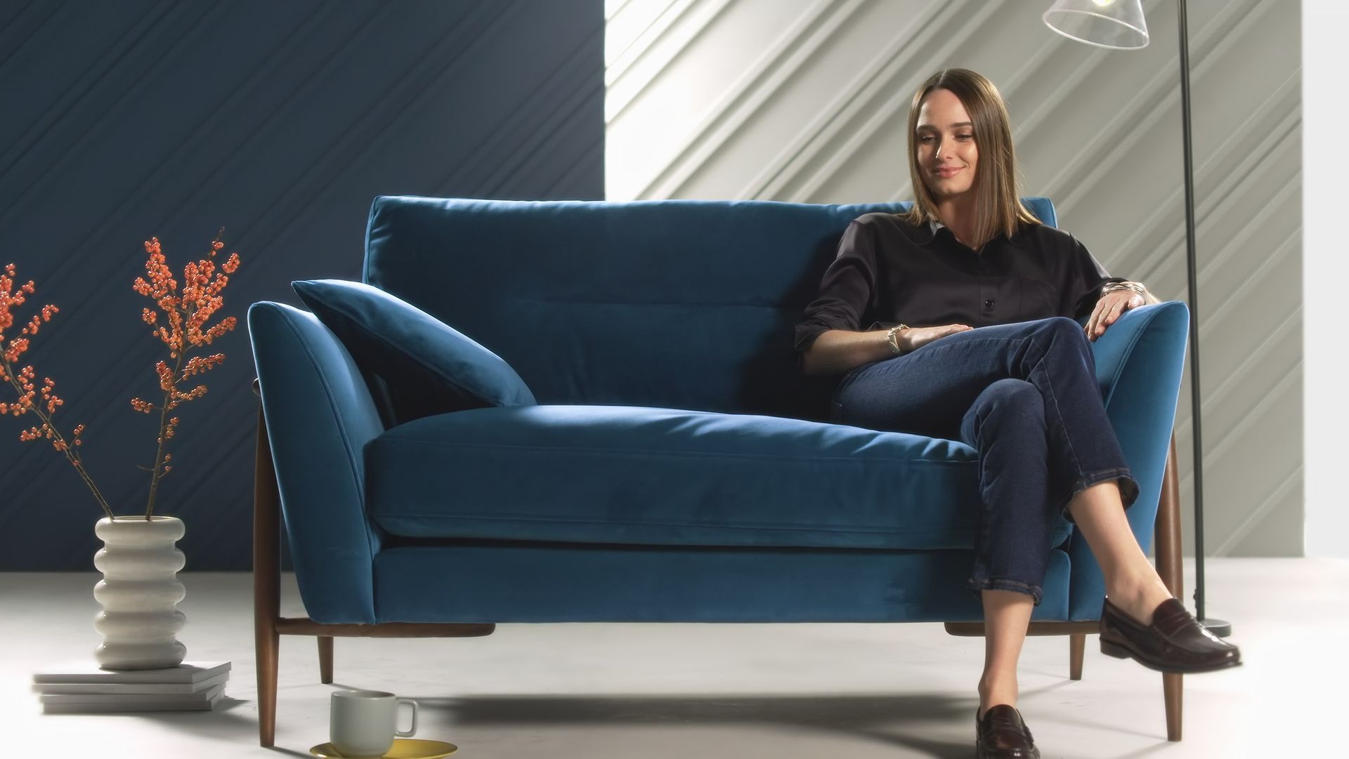 Black Lab Films | A person sits relaxed on a deep blue velvet sofa in a modern studio space with a floor lamp and a small vase nearby.