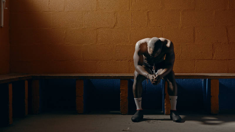 A person sits slumped on a bench in a dimly lit locker room with an orange brick wall.