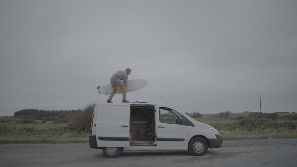 A person in light clothing stands on the roof of a white van, holding a surfboard against a cloudy, rural background.