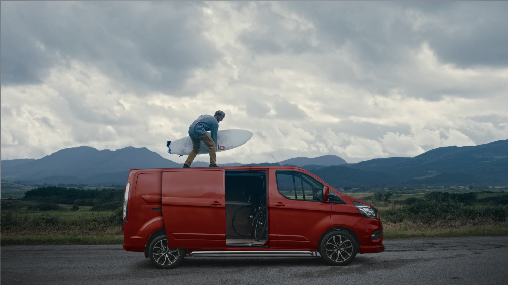 A person stands on the roof of a red van on an open road, holding a surfboard against a backdrop of mountains and sky.