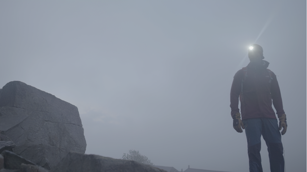 A hiker in a red jacket stands on a rocky, foggy mountainside, wearing a headlamp that shines into the thick mist.