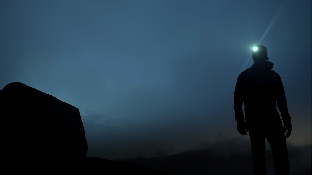 A silhouette of a person wearing a lit headlamp stands outdoors against a dark, moody blue twilight sky.