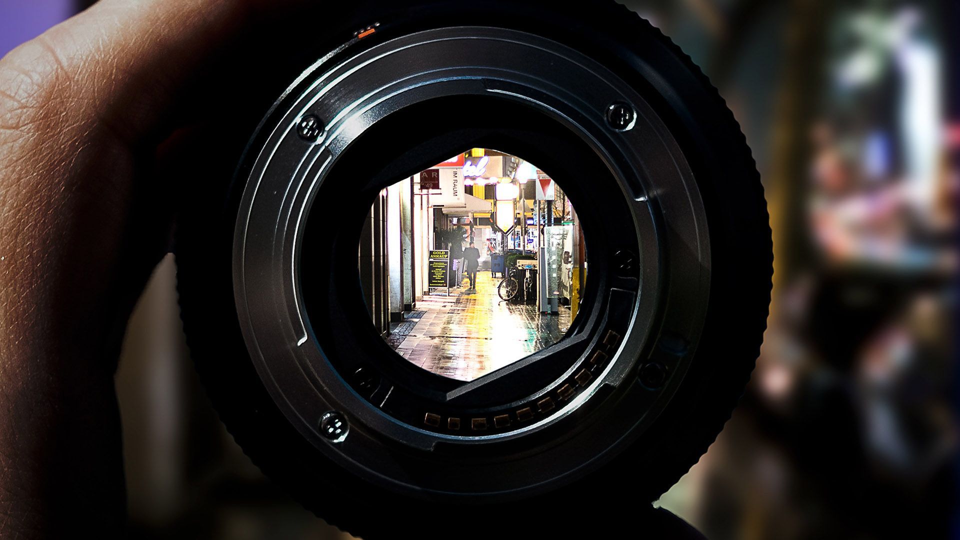 A close-up view through a camera lens, framing a blurry, wet urban street scene at night with warm city lights.