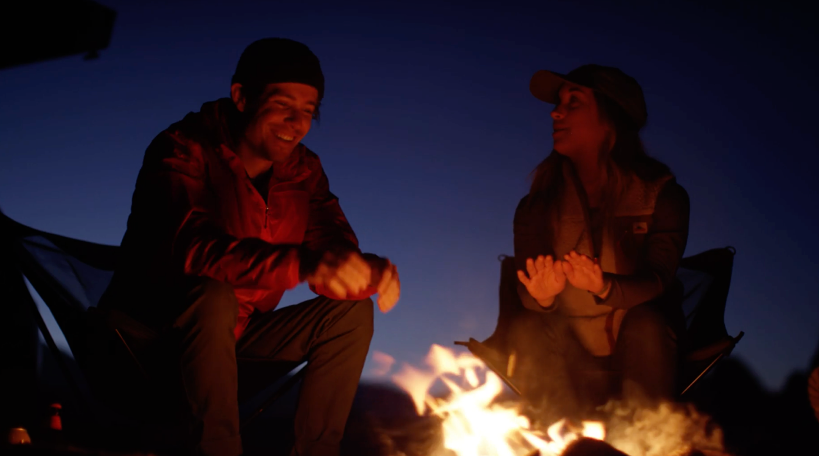Two people sit in camping chairs around a glowing campfire at twilight, warming their hands and talking.