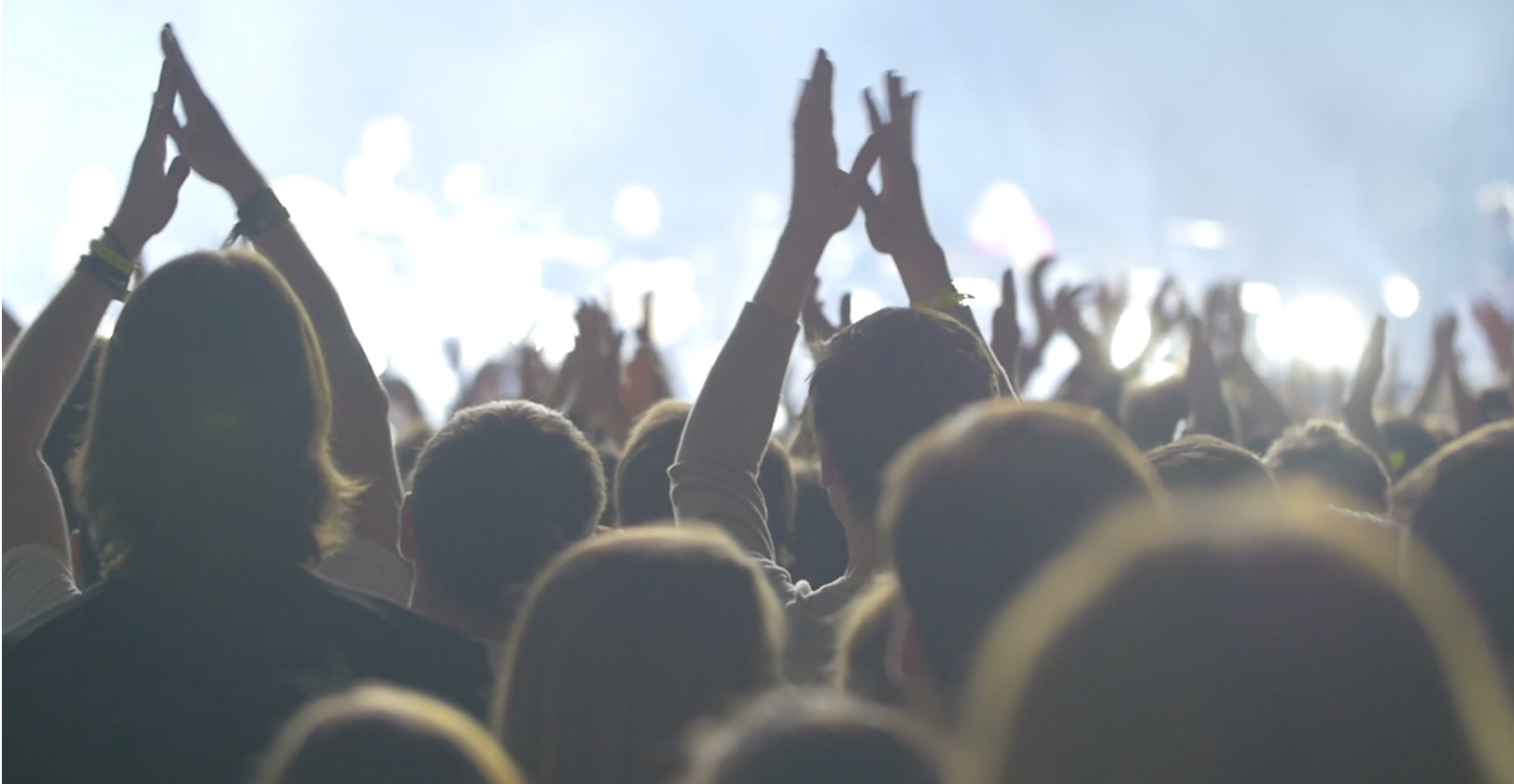 A crowd of people at a concert or event, viewed from behind, with many individuals raising their hands to clap.