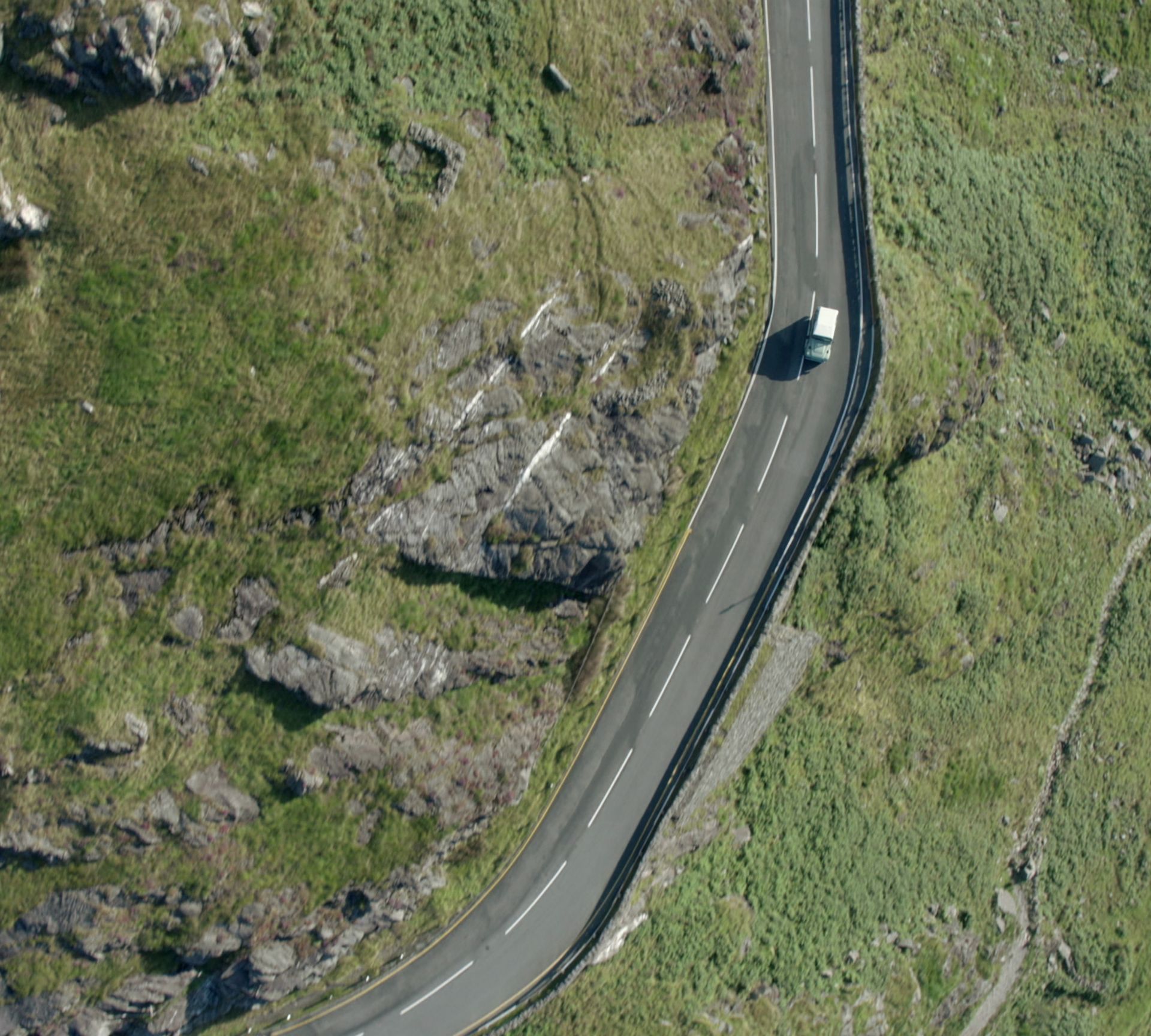An aerial view of a single white vehicle driving along a winding asphalt road through a grassy, rocky mountain landscape.