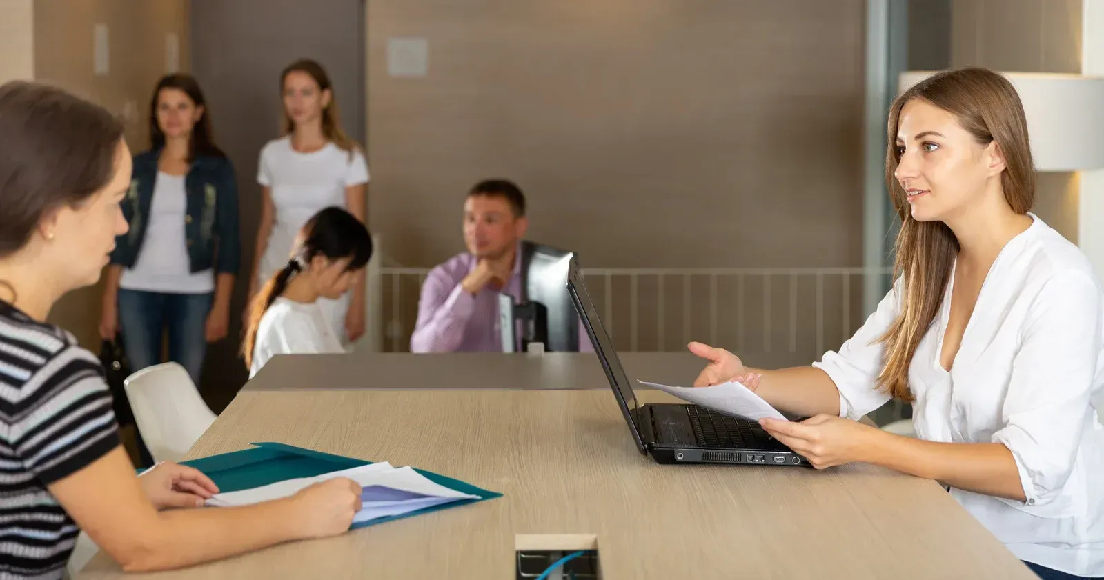 Two women at a table discussing paperwork, a laptop nearby, with other people in the background.