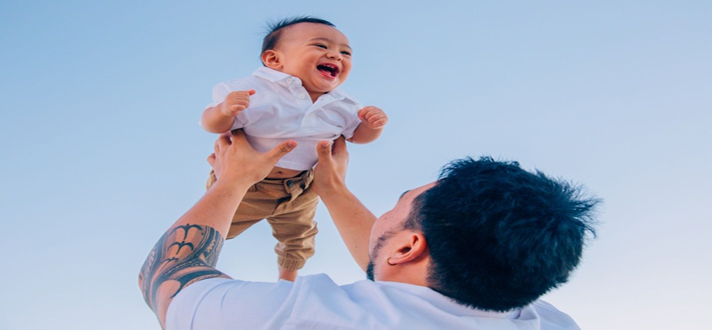 A man is holding his baby in the air with blue sky in the background. The baby is smiling.