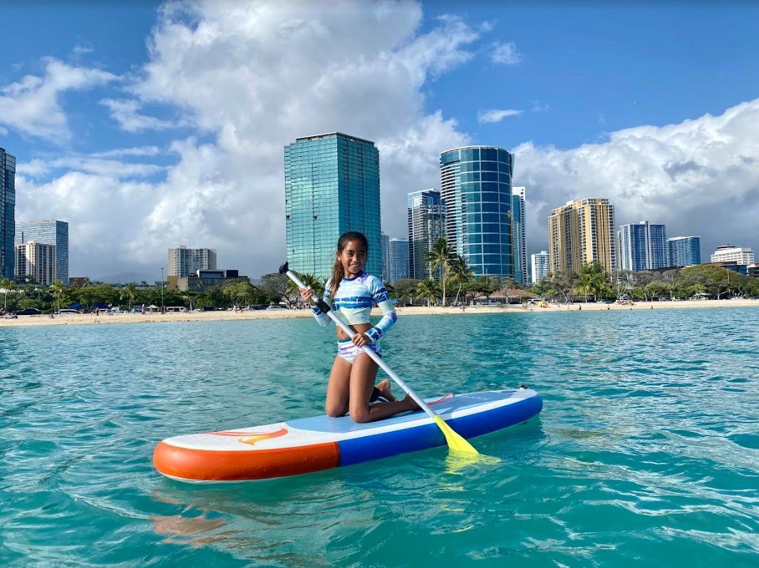 Girl on paddleboard on ocean with tall buildings in the background.