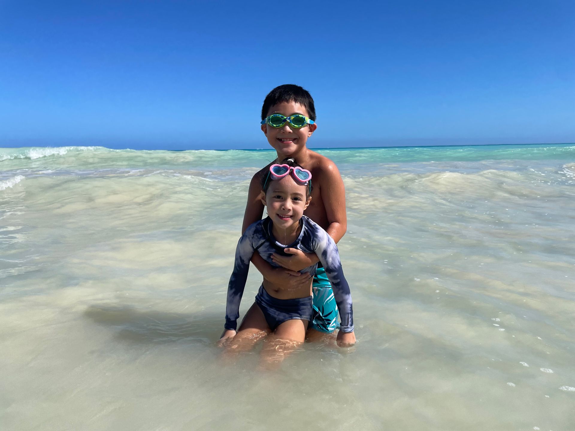 Two children sitting in the water at the beach. They are looking at the cameral and smiling. One is wearing goggles.