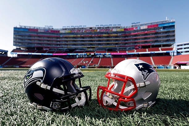 Two football helmets on a field, one navy and one silver with a red facemask, stadium in background