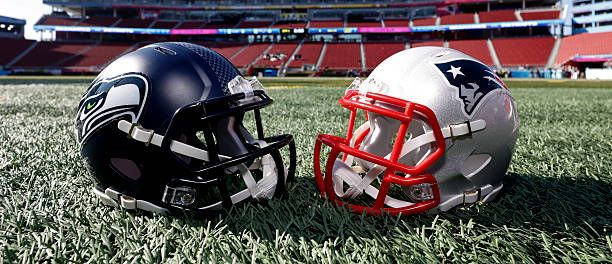 Two football helmets on a field in front of a stadium, navy and silver with red facemasks