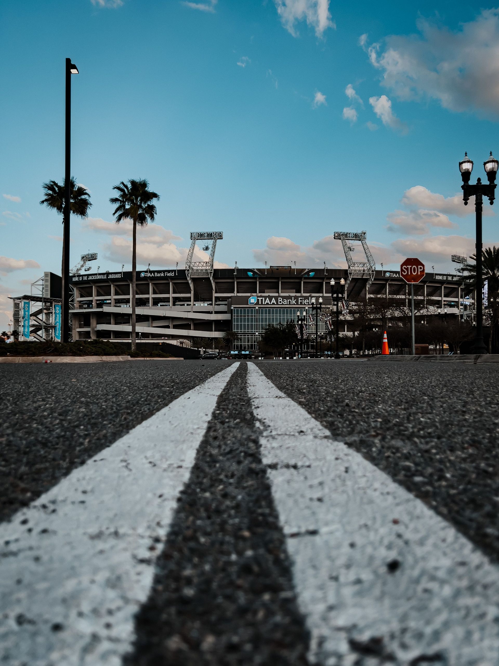 Low-angle view of an empty road leading to a stadium under a blue sky with palm trees
