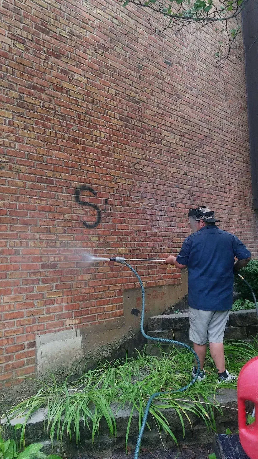 Person in a blue jacket spray-paints a brick wall near a garden hose and plants.