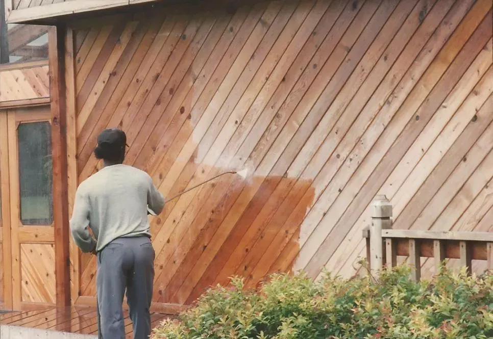 Person staining a wooden wall with a large brush outdoors.