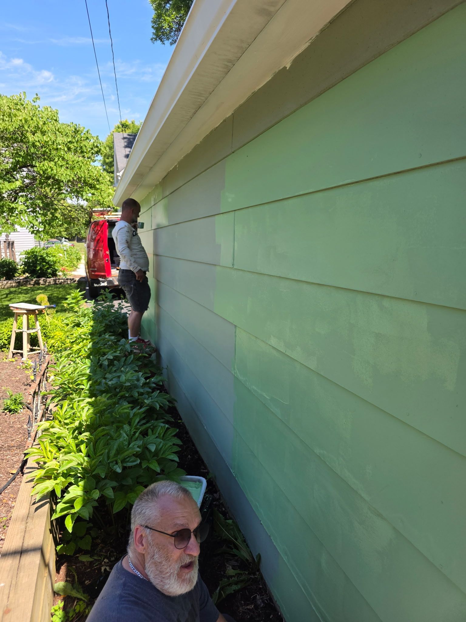 People standing beside a green house wall along a narrow garden path in bright sunlight