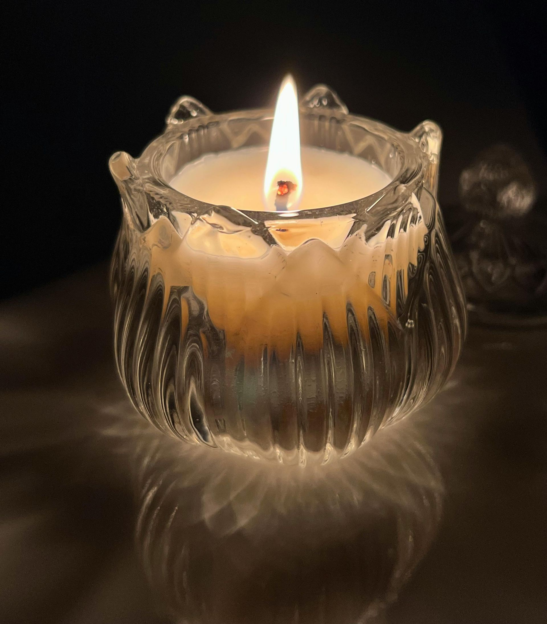 Burning candle in a clear, decorative glass container casting light shadows on a dark surface.