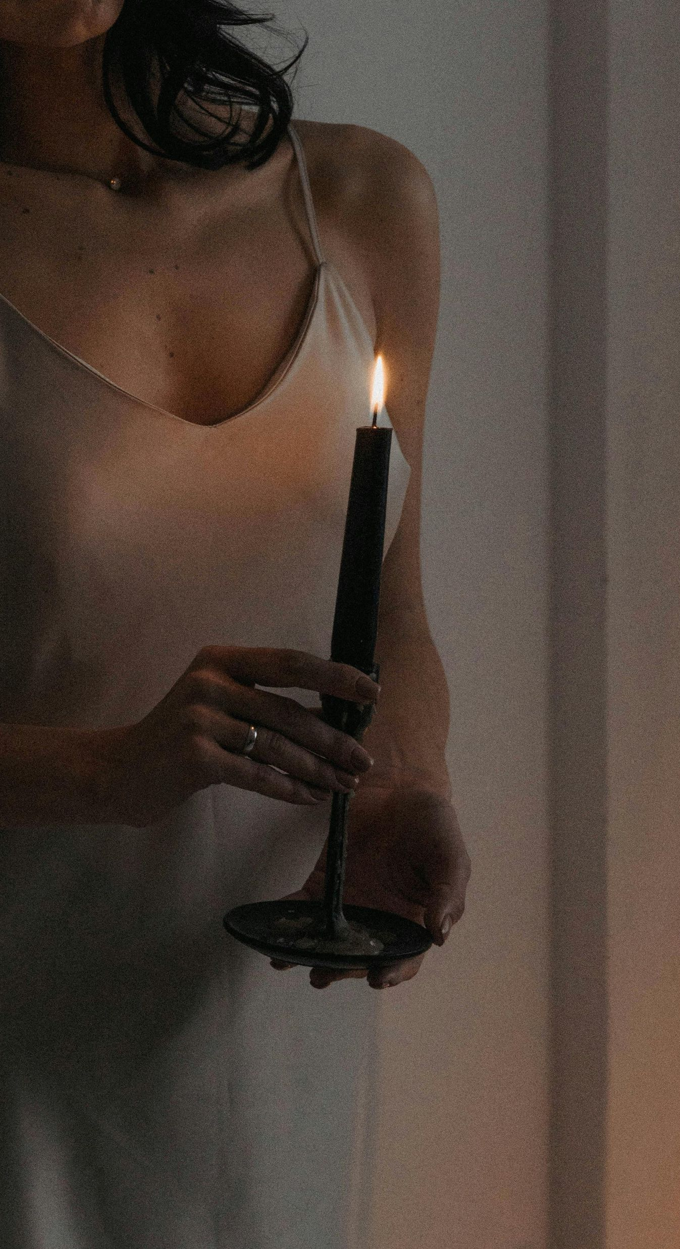Woman holding a lit black candle, wearing a light-colored dress in a dim room.