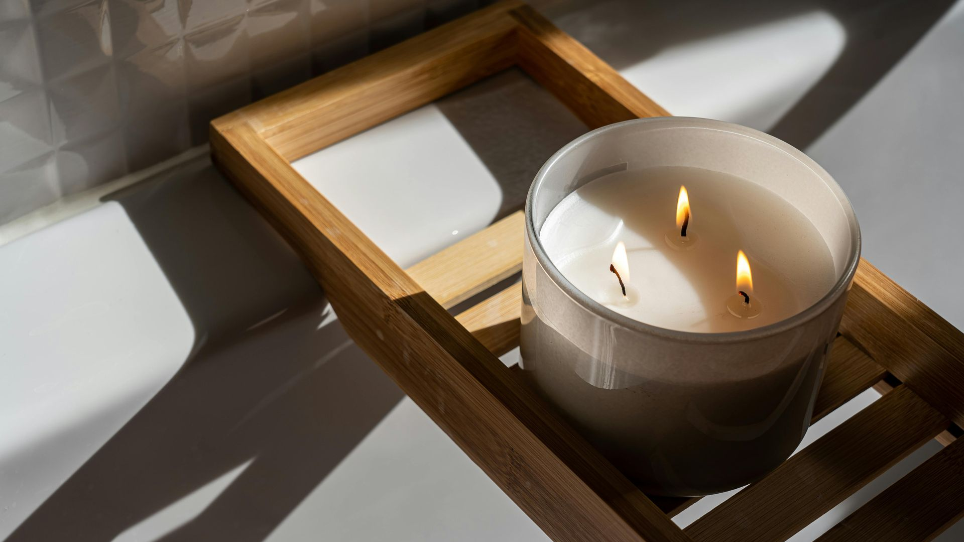 Lit candle in a clear glass jar on a wooden bath tray, casting shadows against a white tile background.
