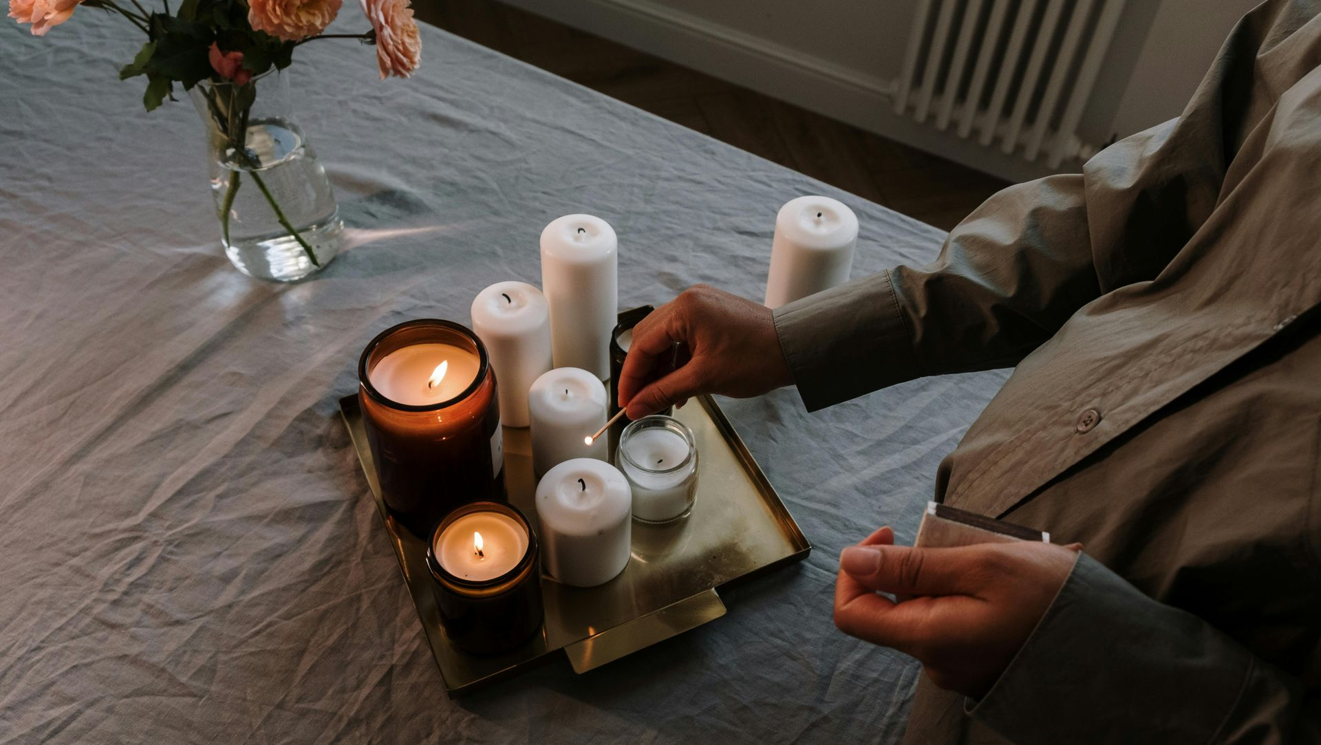 Person lighting candles on a tray on a table, next to a vase of flowers.