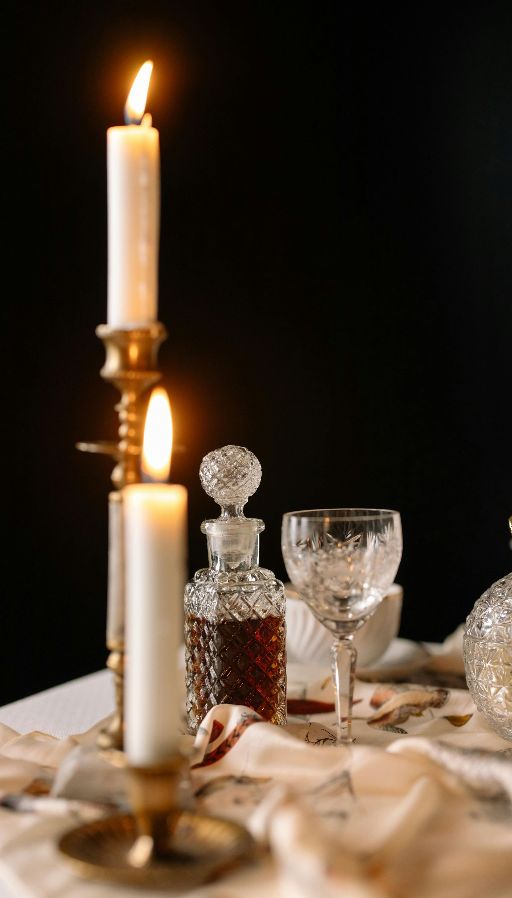 Lit candles, crystal decanter, and glass on a table with dark background.