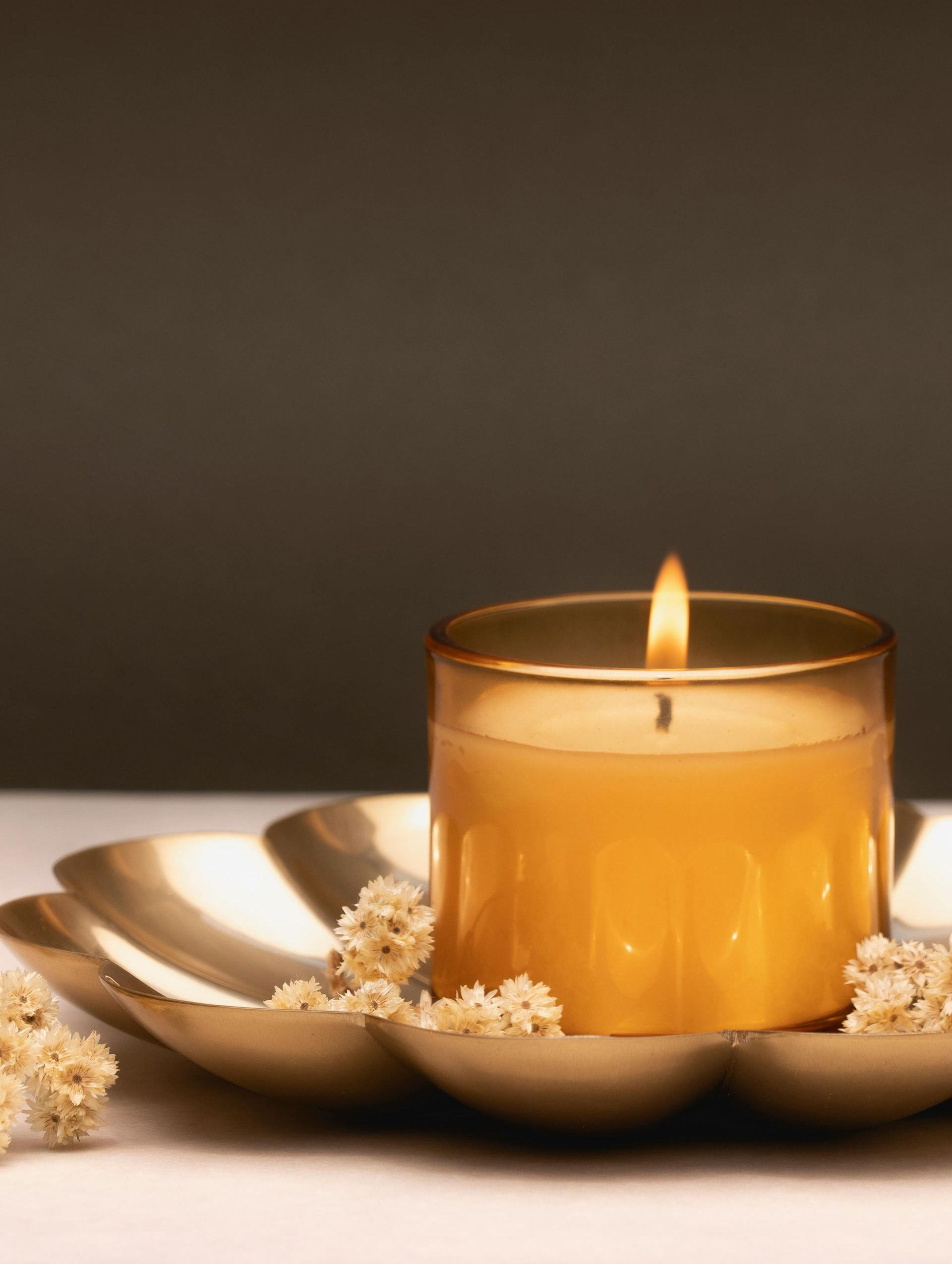Lit yellow candle in a gold glass jar, on a gold decorative tray with white flowers; neutral background.