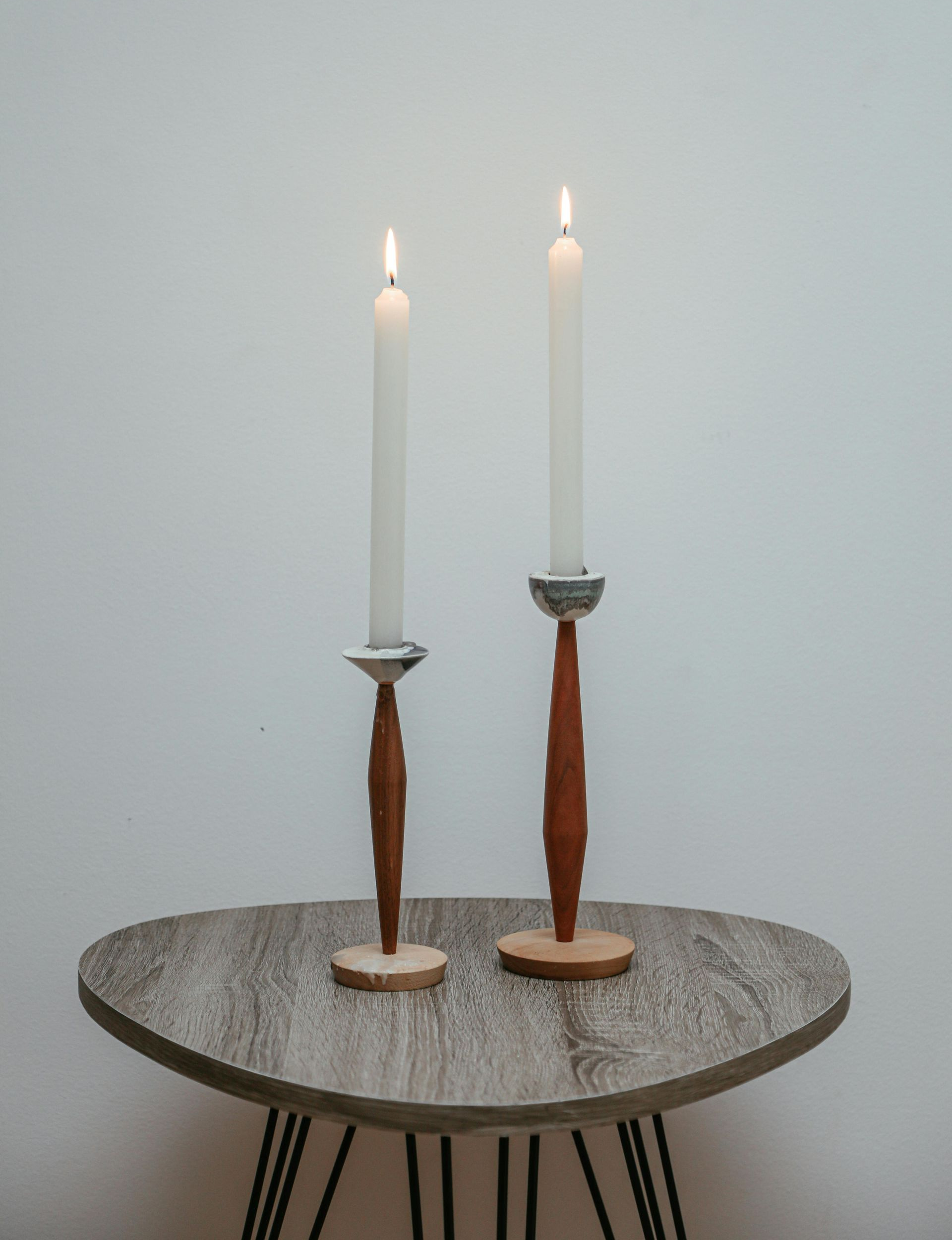 Two lit candles on wooden candlesticks atop a textured table against a white wall.