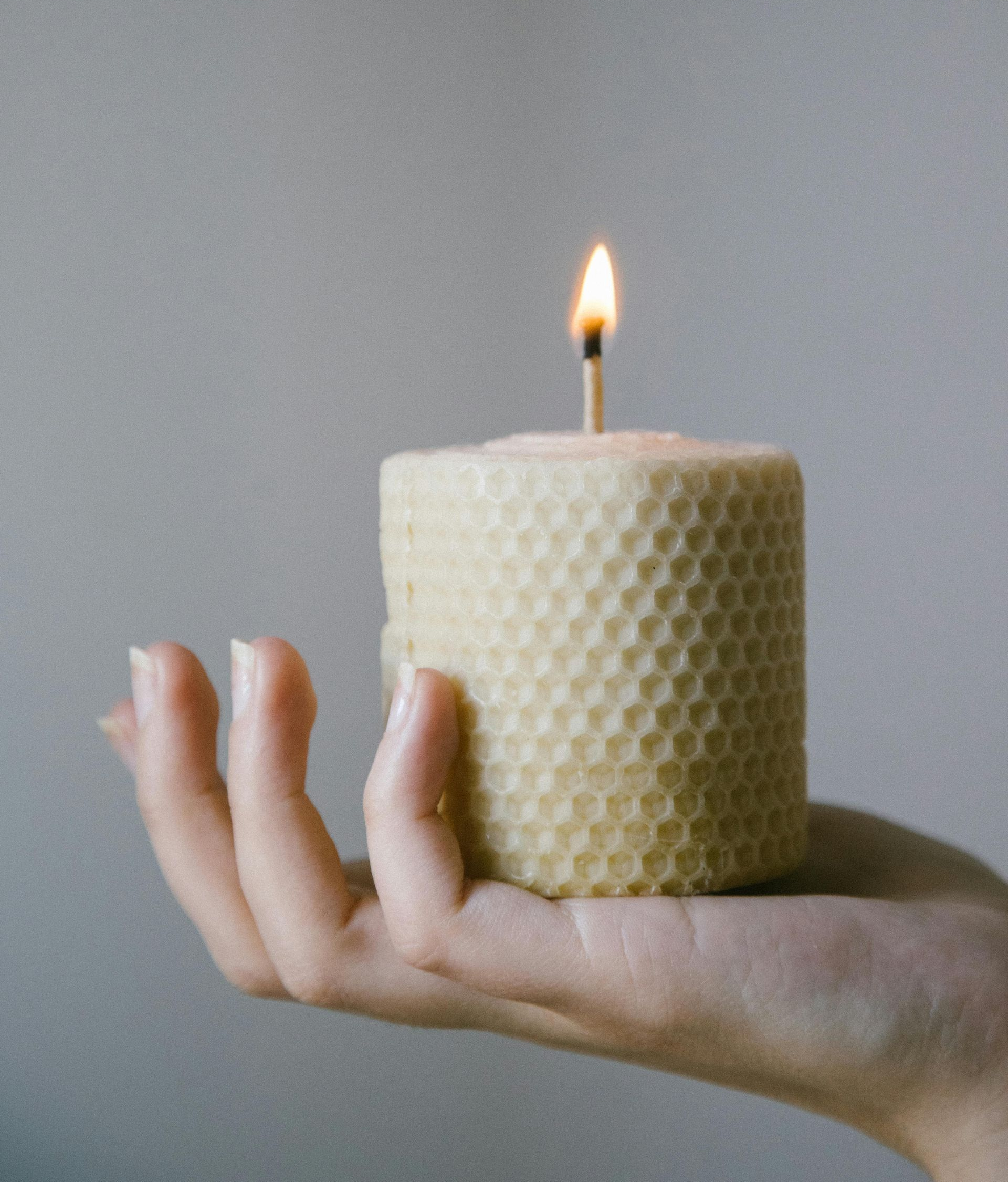 Hand holding a lit, cylindrical beeswax candle with a honeycomb pattern; neutral background.