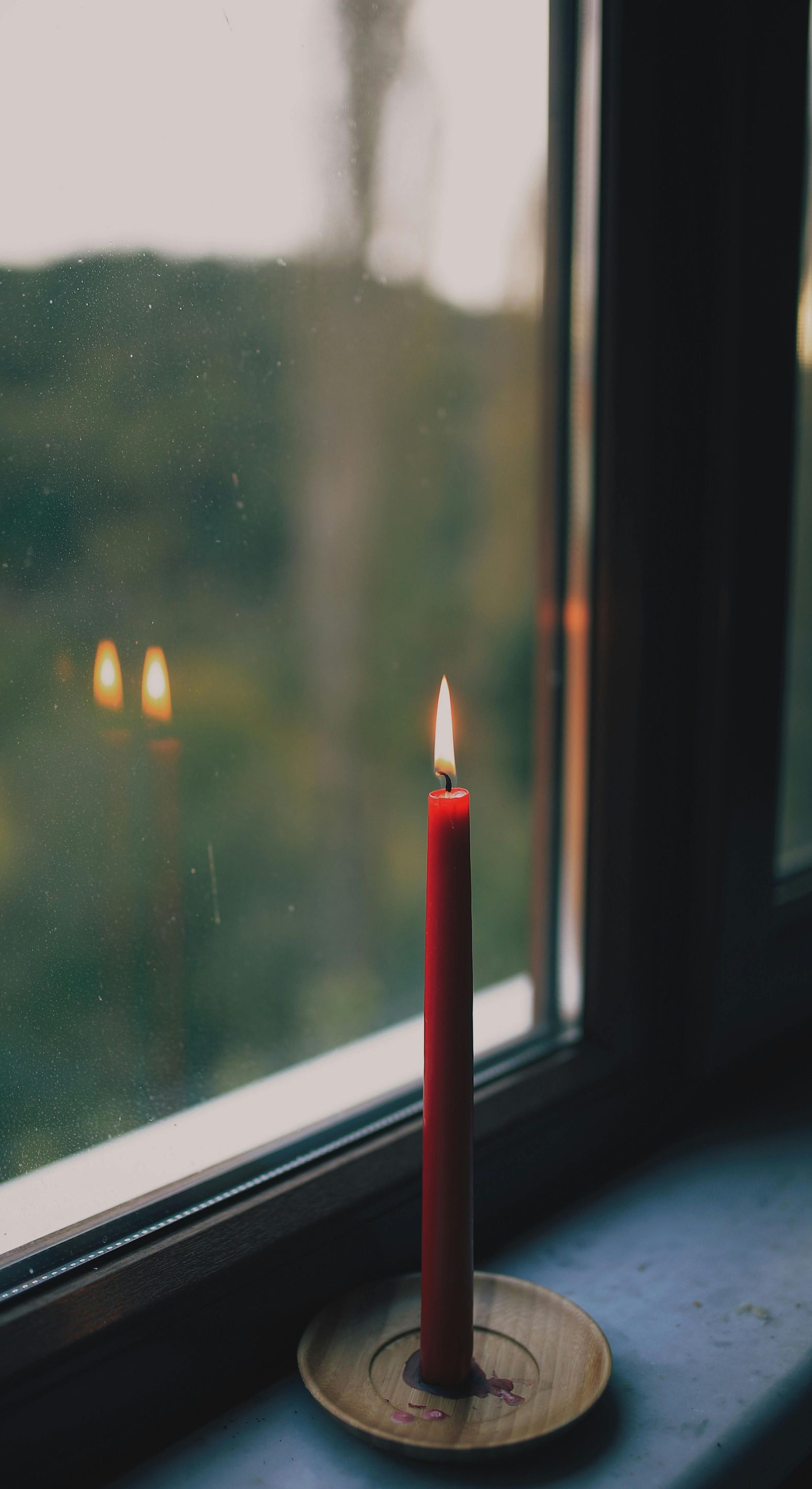 Red candle burning on a brass holder, placed on a windowsill. Outside, a blurry green view.