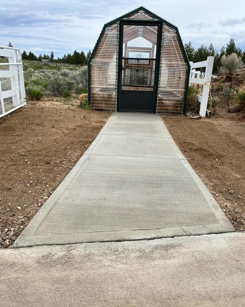 Concrete path leads to a wooden structure with a dark door, nestled between white fences and the outdoors.