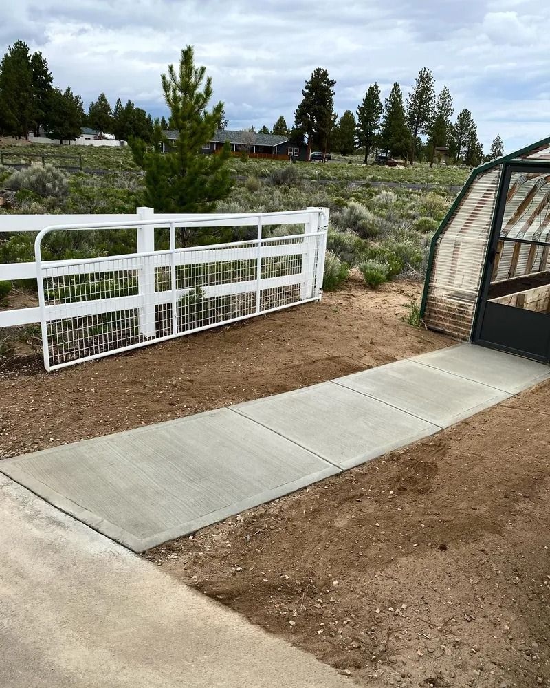 Concrete walkway leading to a white fence and a greenhouse. Outdoors, cloudy sky, dirt ground.