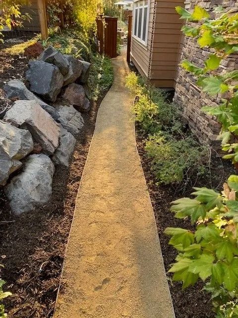 A gravel path winds between rocks, a house, and greenery. Sunlight illuminates the scene.