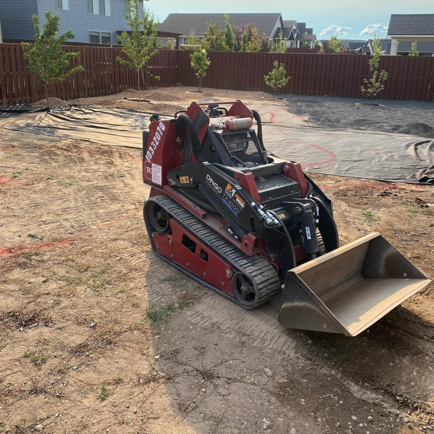 Red compact track loader with bucket on dirt, in a backyard. Brown fence and buildings in background.