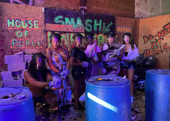 People posing in a neon-lit smash room with barrels and signs reading “House of Purge” and “Do Not Paint Walls”
