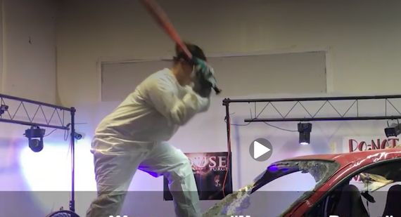 Person in white protective suit swinging a bat and hitting a red car in an indoor event space at house of purge