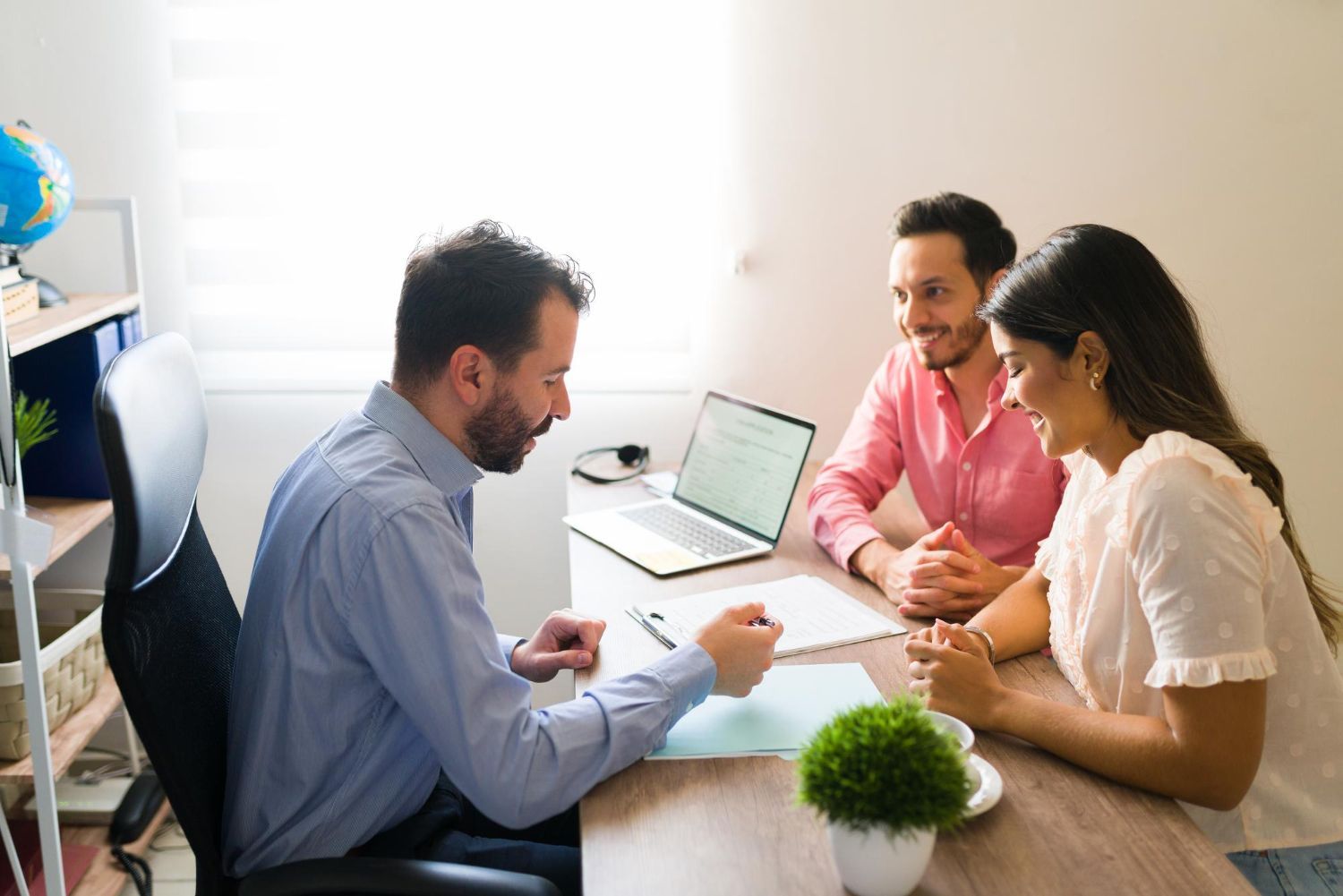 A man in a blue shirt reviews documents with a smiling couple at a desk in an office.