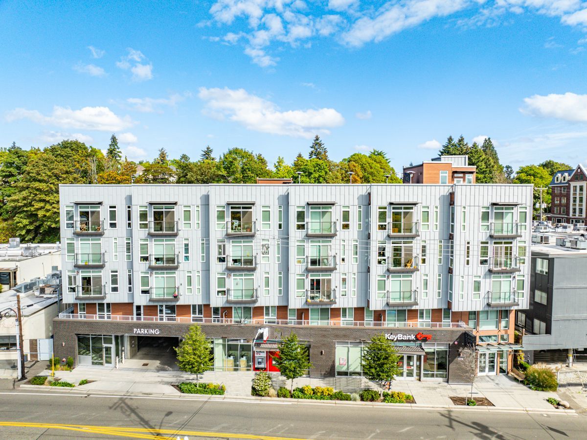 Modern white apartment building with balconies, street-level shops, and trees under a blue sky