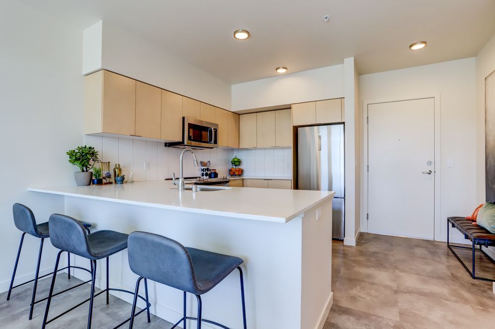 Modern kitchen with white counters, light wood cabinets, stainless fridge, and blue bar stools