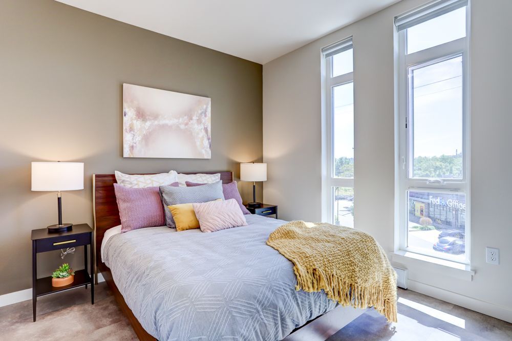 Bright bedroom with a gray bed, pink pillows, bedside lamps, and tall windows with sunlight.