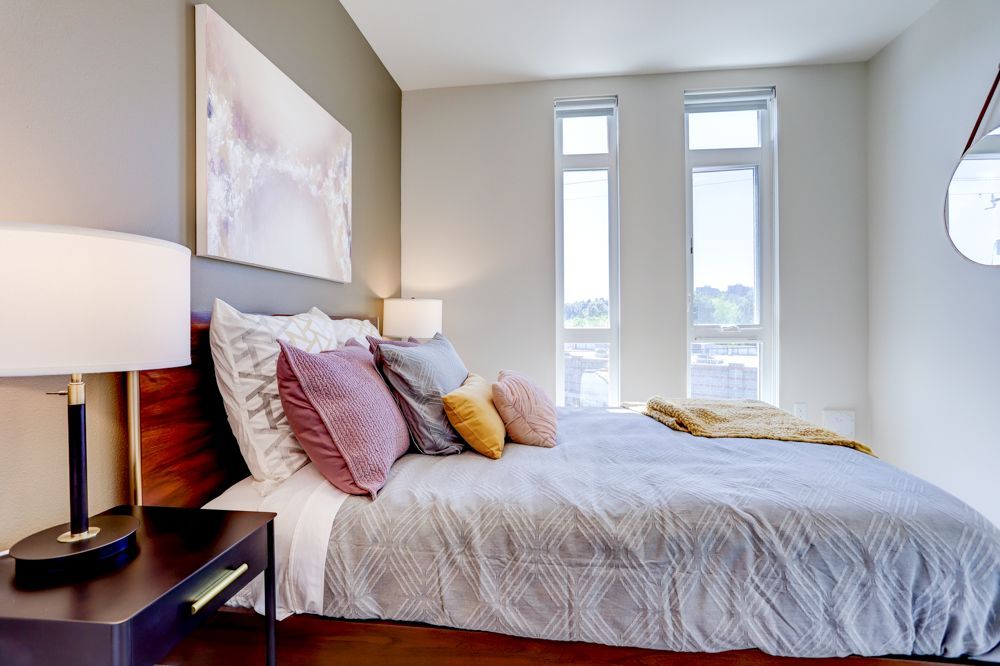 Modern bedroom with a made bed, colorful pillows, bedside lamps, and tall windows.