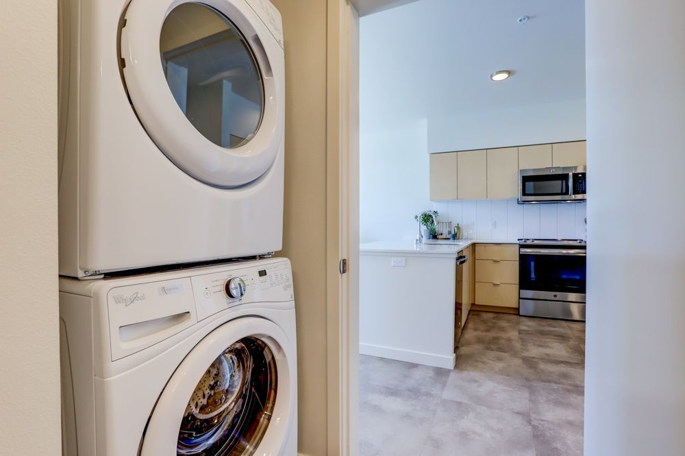 Stacked washer and dryer in a hallway, with a view into a bright kitchen beyond.