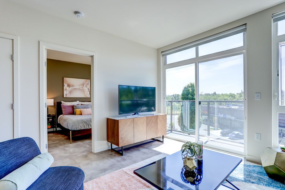 Modern living room with blue sofa, TV, coffee table, and floor-to-ceiling windows overlooking a balcony.