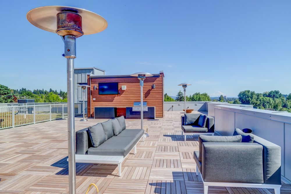 Sunny rooftop lounge with gray seating, patio heaters, and a small wooden building by the railing