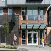 Modern apartment building entrance with glass canopy; two pedestrians pass by on the sidewalk.