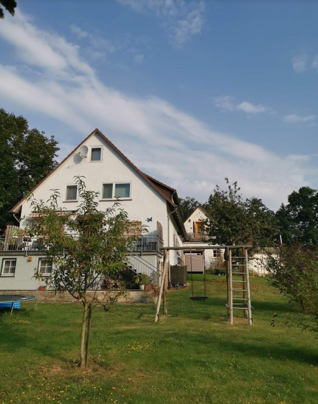 Wit huis met een schuin dak, schommel ervoor, groen gazon, blauwe lucht.