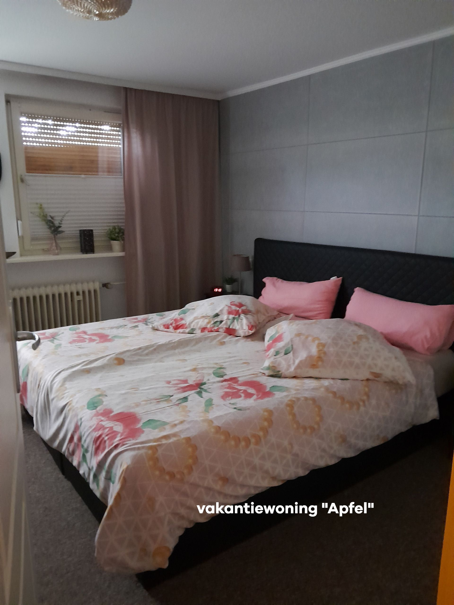 Slaapkamer met een groot bed, roze kussens, beddengoed met bloemenprint en een raam met jaloezieën.