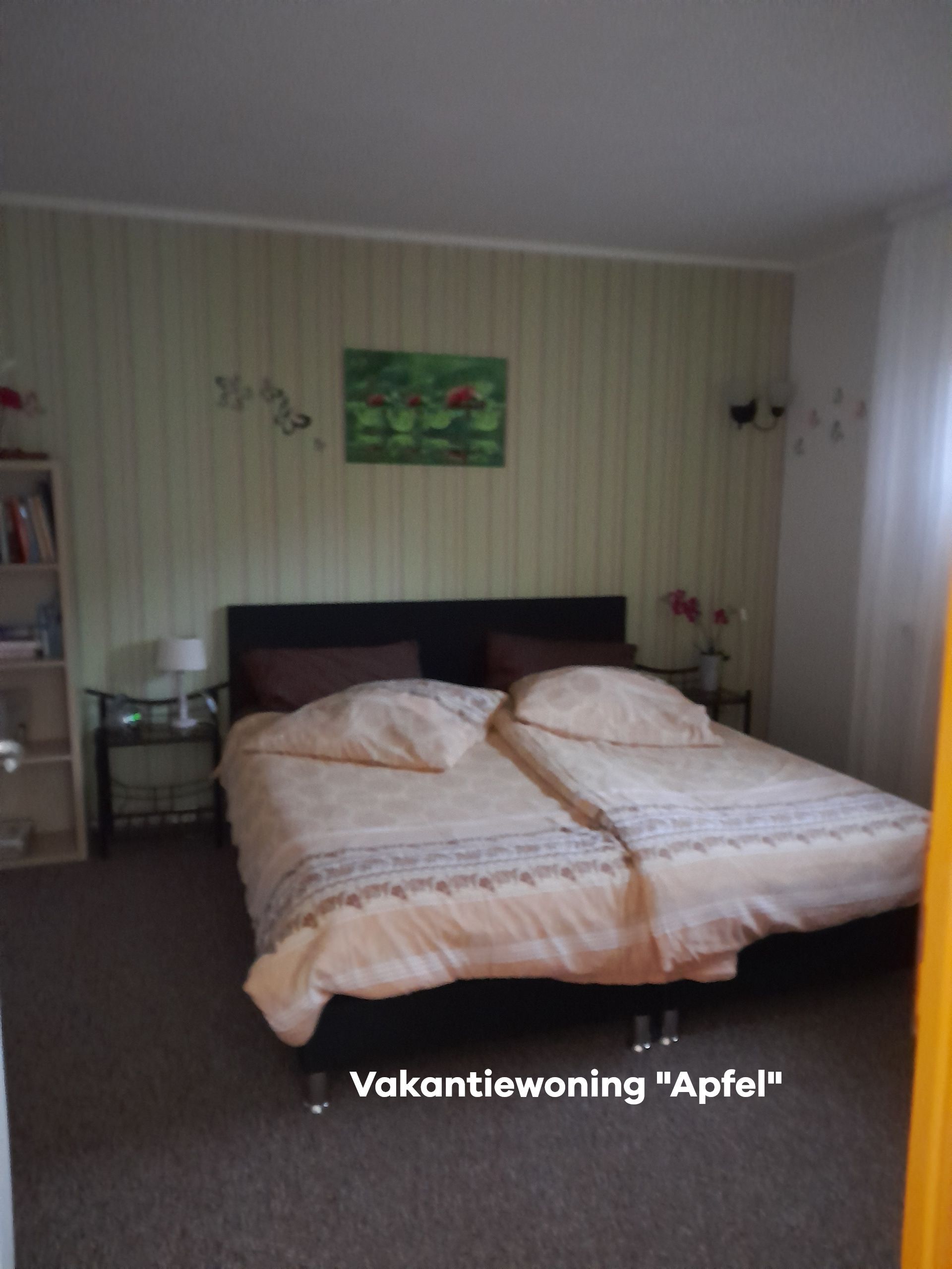 Slaapkamer met twee eenpersoonsbedden, beige beddengoed en een zwart hoofdeinde. Groen gestreepte muren, kunst, een nachtkastje en een boekenkast.